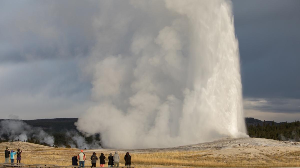 Old Faithful erupting under a stormy sky as visitors watch from the boardwalk