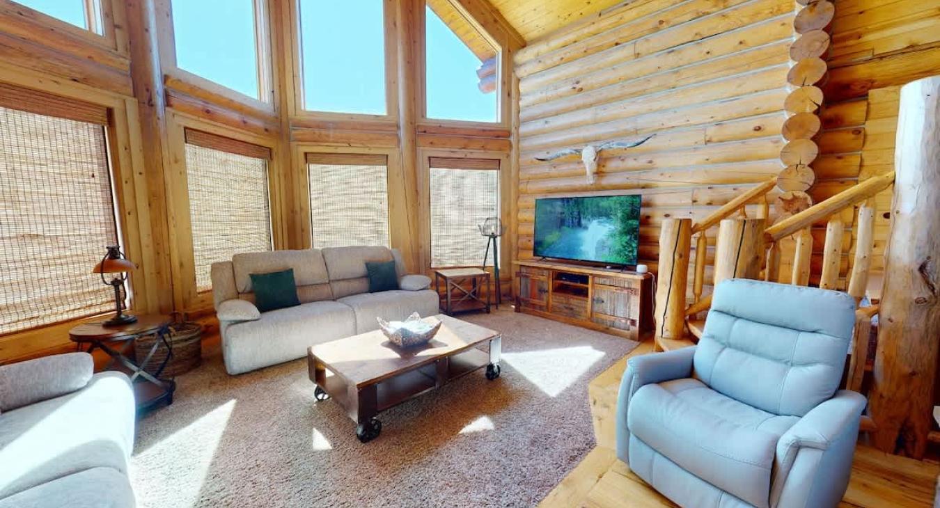 The room is well-lit by natural light pouring in from large, floor-to-ceiling windows. A flat-screen television is placed on the entertainment center, offering modern convenience without detracting from the cabin’s rustic appeal.