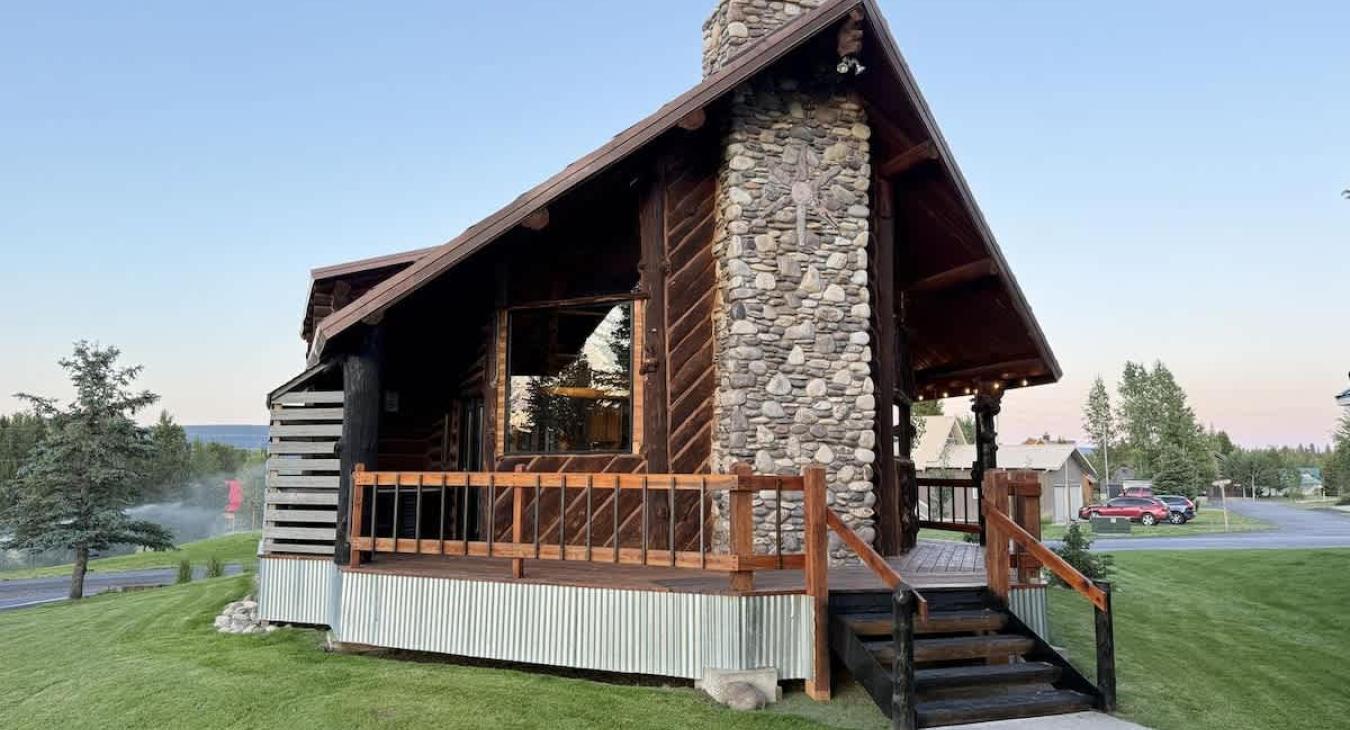 The cabin's exterior is a harmonious mix of natural materials, with large logs and weathered wood giving it an authentic, mountain cabin feel.