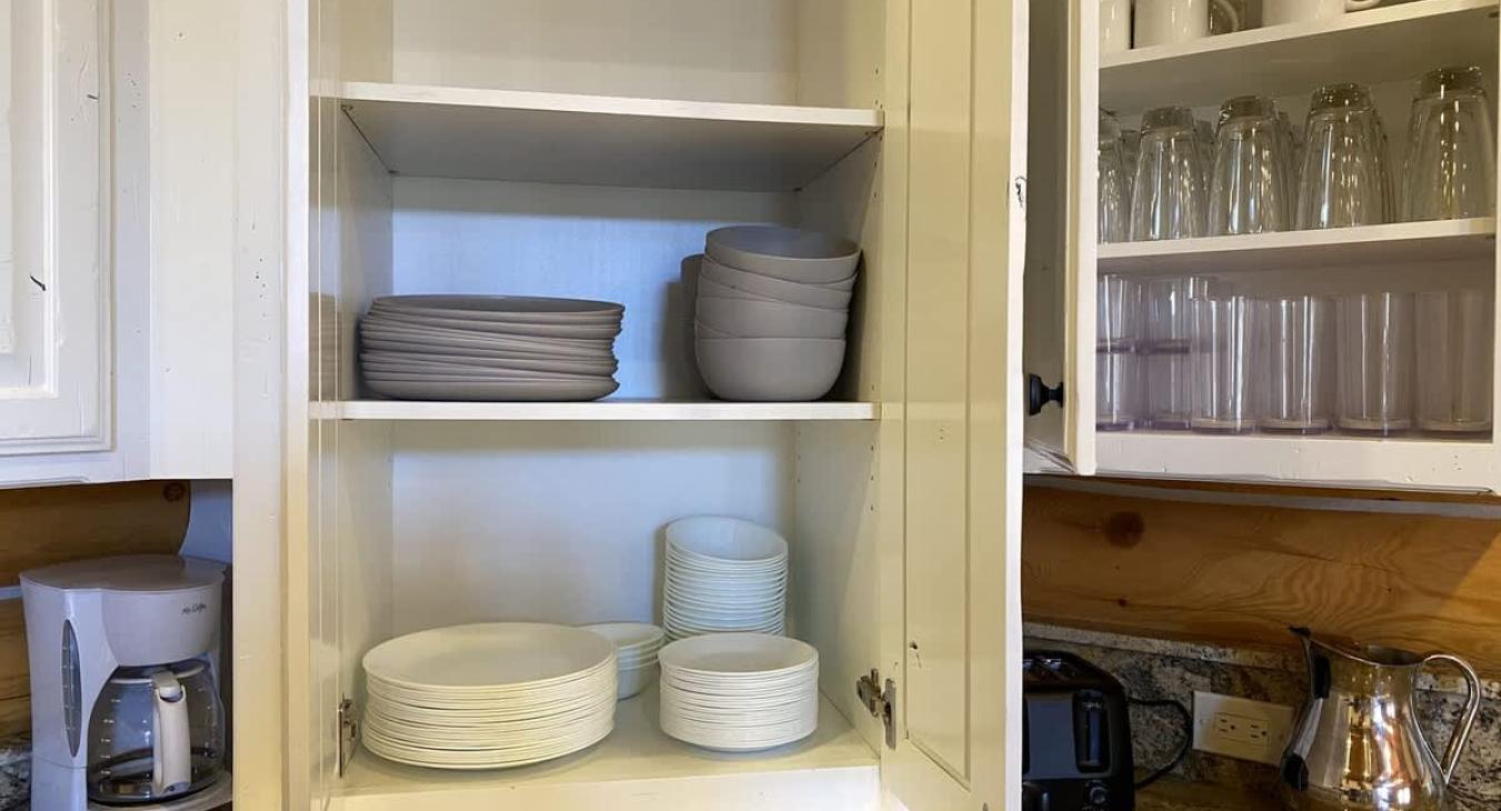 The white kitchen cabinets have a fresh, clean look, offering a modern yet cozy contrast to the rustic charm of the cabin. Neatly arranged, you’ll find a set of glass cups, mugs, plates, and bowls.