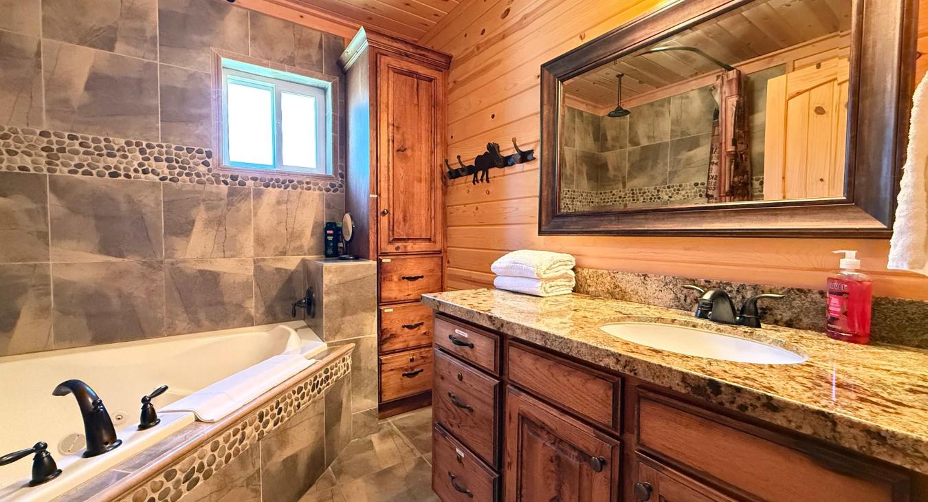 The luxury en-suite master bathroom boasts a large custom wooden vanity with a premium granite countertop, bright lighting, and a deep soaking jetted tub.