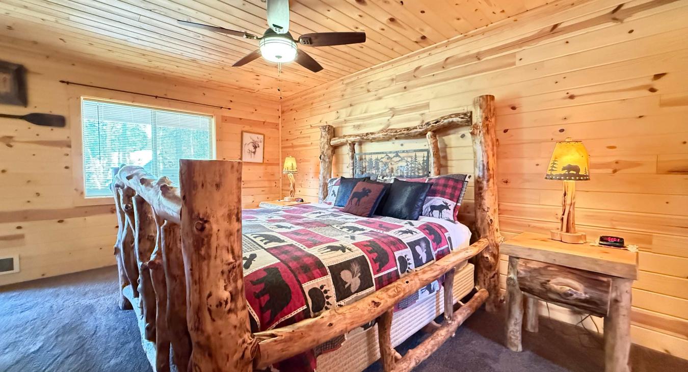 The beautiful master bedroom features an authentic rustic log-framed King bed, a cozy ceiling fan, and warm wood-paneled walls that create a perfect mountain cabin retreat.